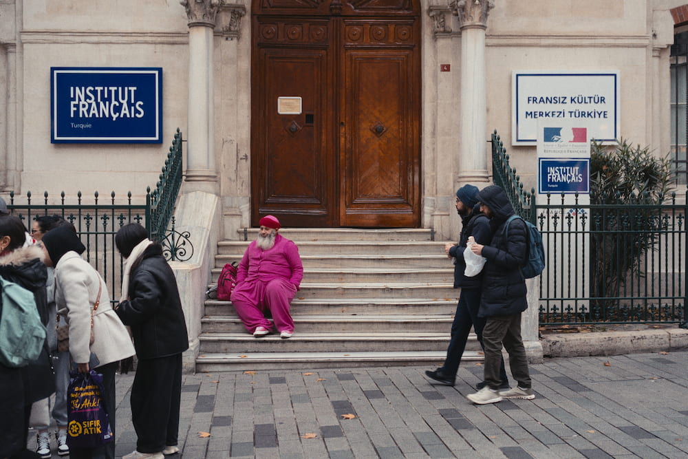 A man in pink sitting on the steps of the Institut Français, people passing.