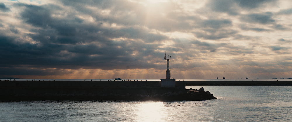 A breakwater and a small light tower against sun breaking through cloud.