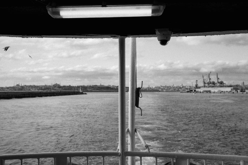 The view aft from under a ferry roof, cranes on the far shore, in black and white.