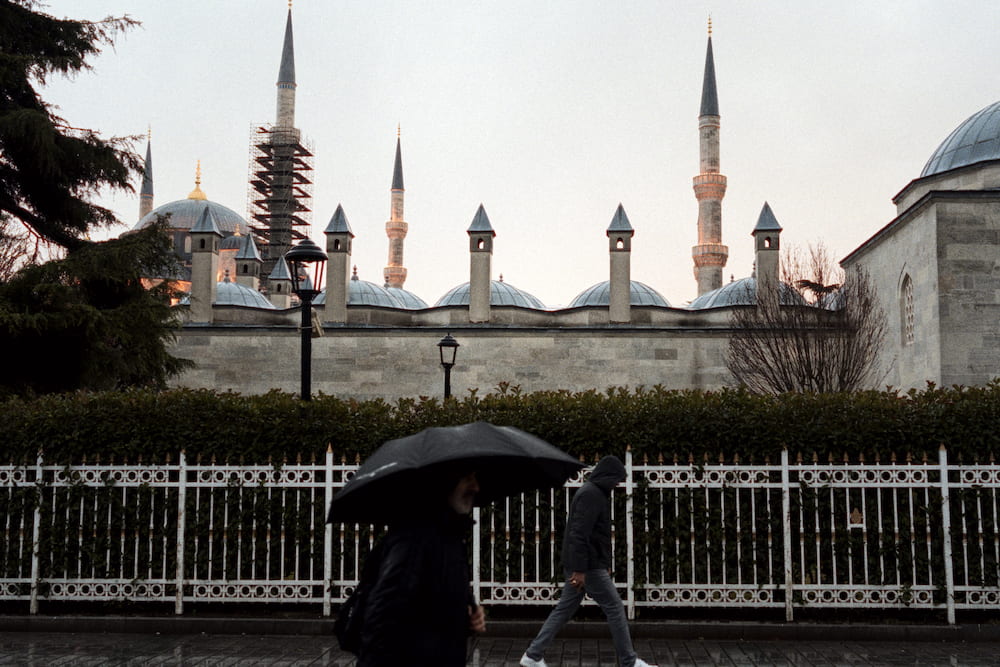 A man with an umbrella walking past the domes and minarets of a mosque.