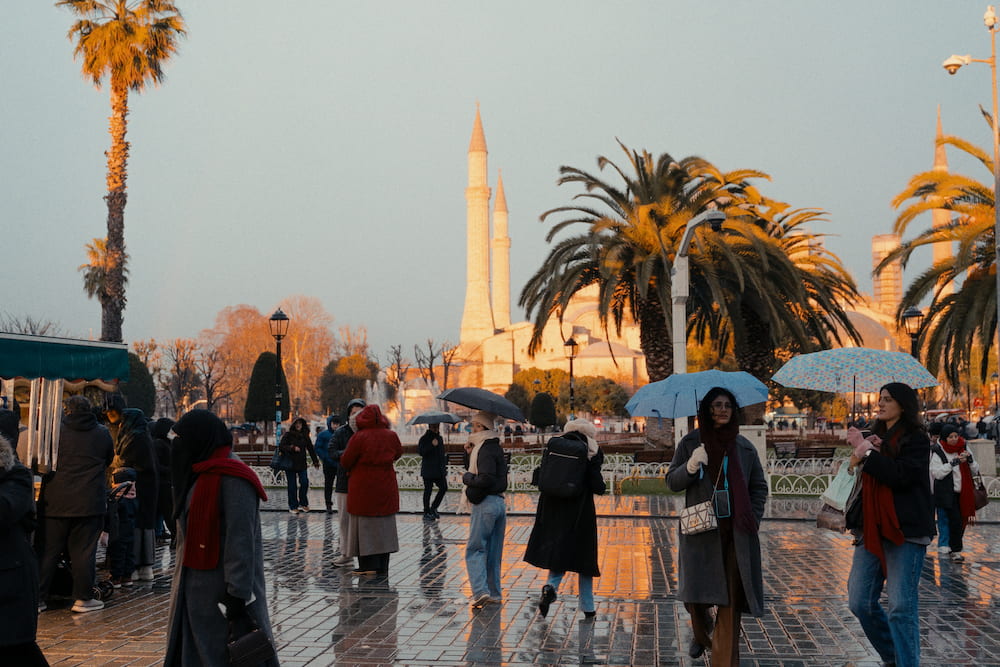 People with umbrellas on a wet square in low sun, palm trees and minarets behind.