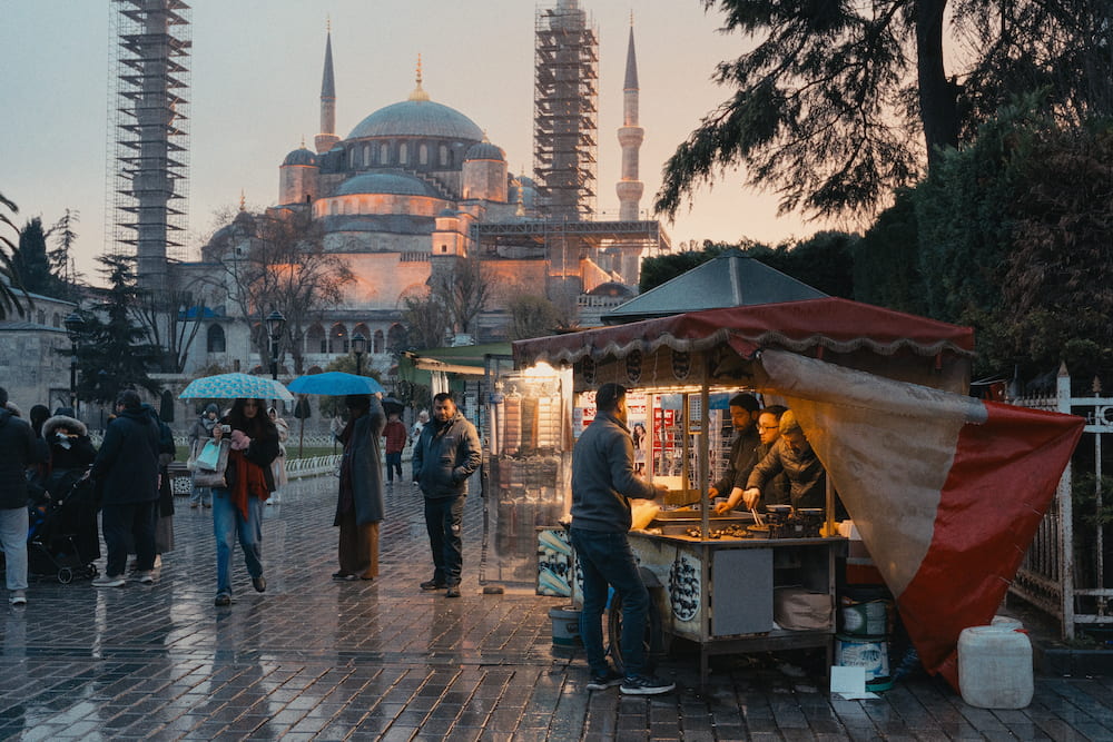 A street food cart with a red awning on wet paving, a mosque and two minarets in scaffolding behind.