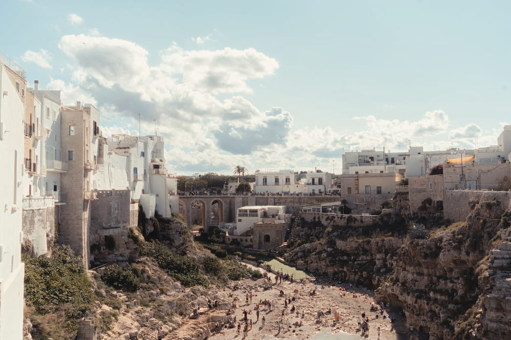 Sunbathers in a small cove below limestone cliffs, with white houses and an arched bridge above