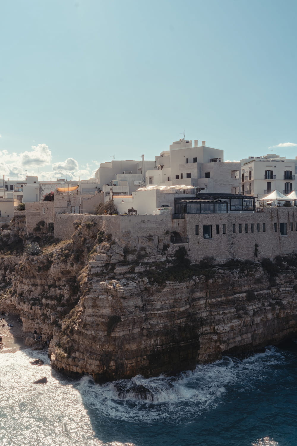 White buildings along the top of a layered limestone cliff, waves breaking at its foot