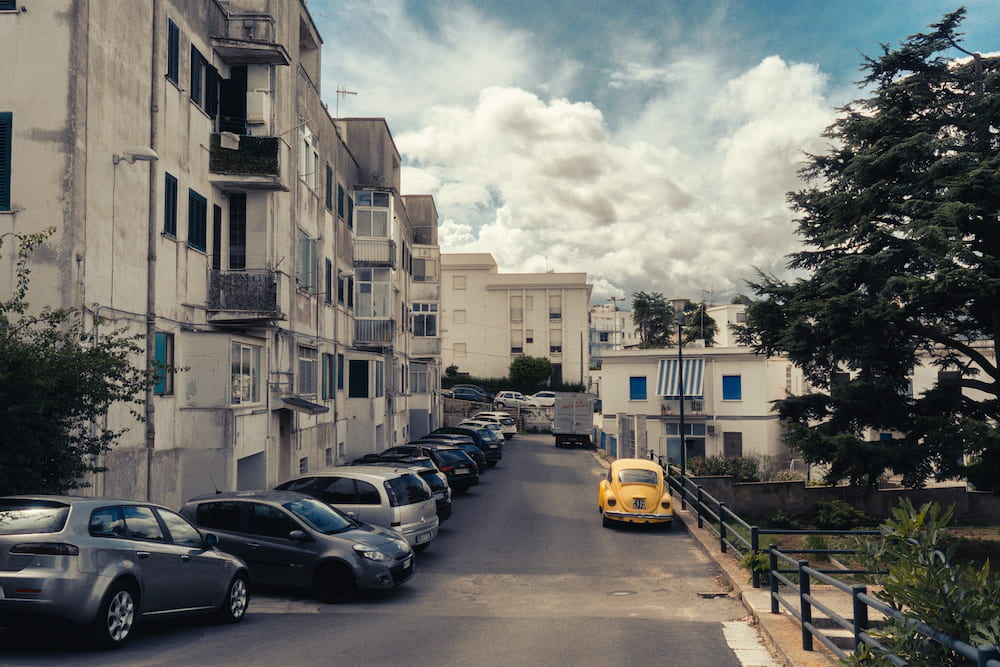 Yellow Beetle parked on a quiet street of apartment blocks and parked cars under big clouds