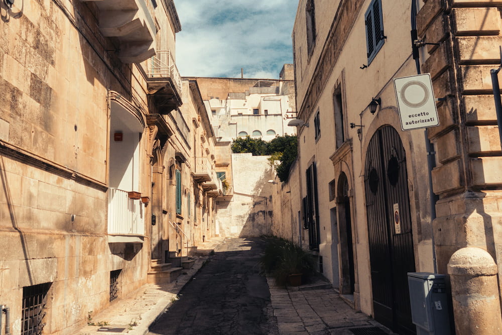 Narrow lane of pale stone houses with balconies and dark arched doors, sunlit at the far end