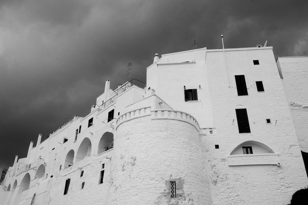Black and white low view of whitewashed arches and a round tower under a dark stormy sky