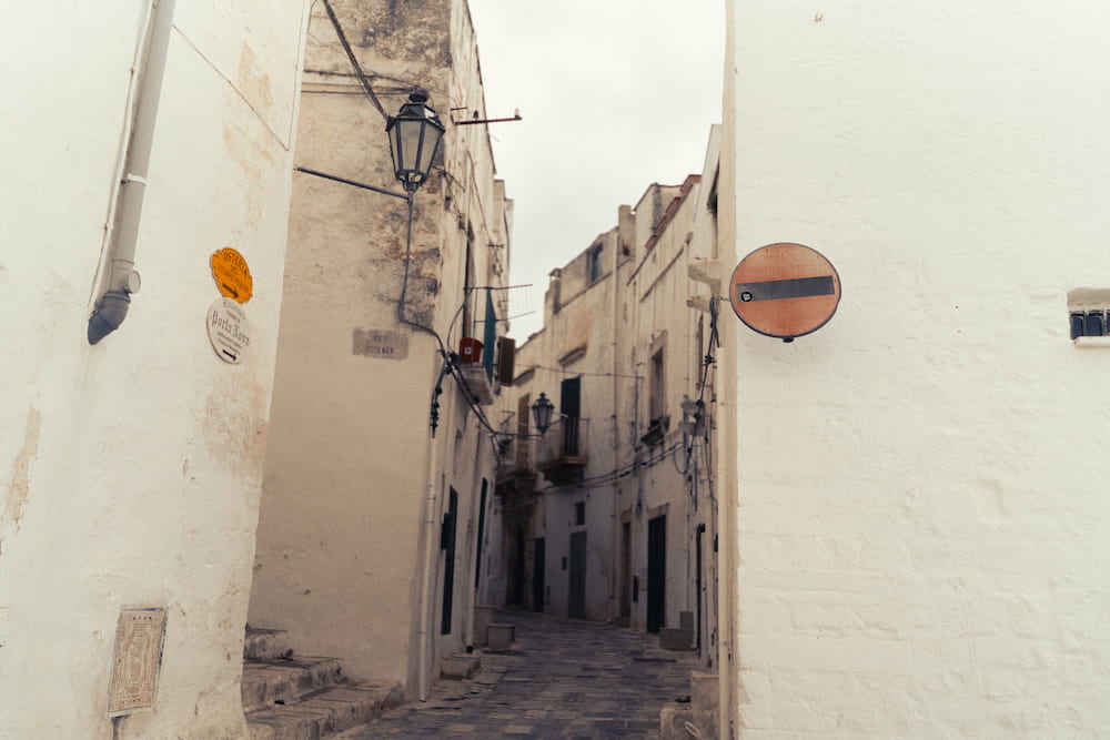 Narrow stone lane between whitewashed walls with old street lamps and a no-entry sign, overcast light