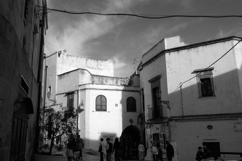 Black and white small square of whitewashed buildings with an arched passage and people under a cloudy sky