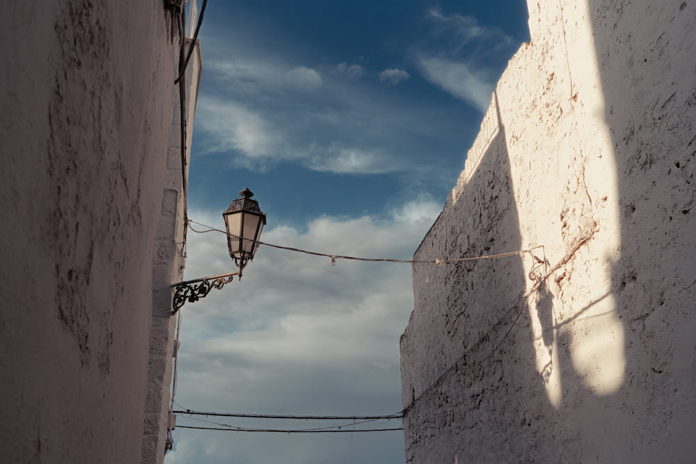 Old street lamp on an iron bracket between two whitewashed walls, one lit by a strip of sun