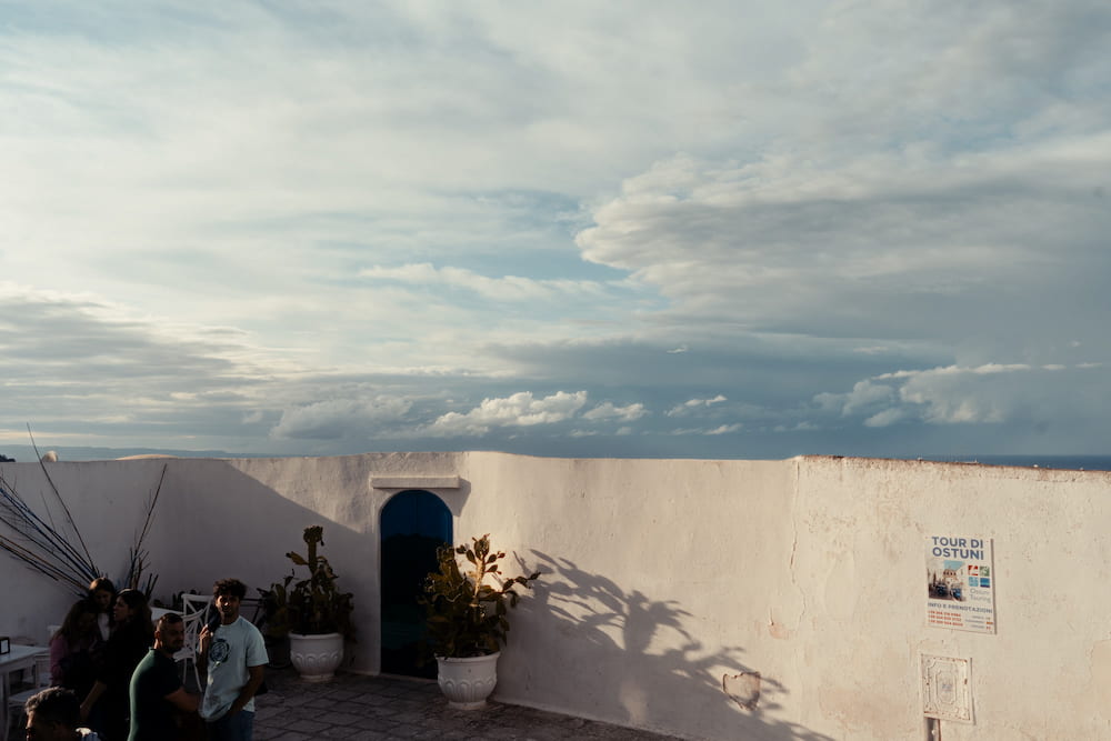 Rooftop terrace with a whitewashed parapet, potted plants, a blue arched door and people under wide clouds