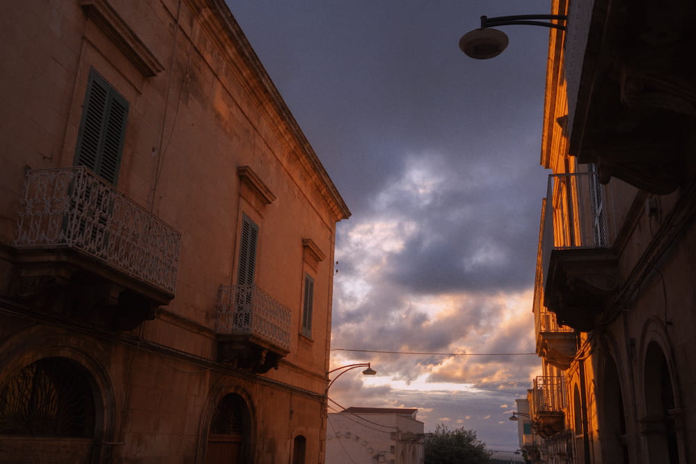 Street of stone buildings with iron balconies against a cloudy sky lit orange at sunset