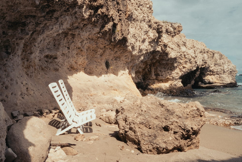 Two white folding chairs on sand under an overhanging limestone rock beside the sea