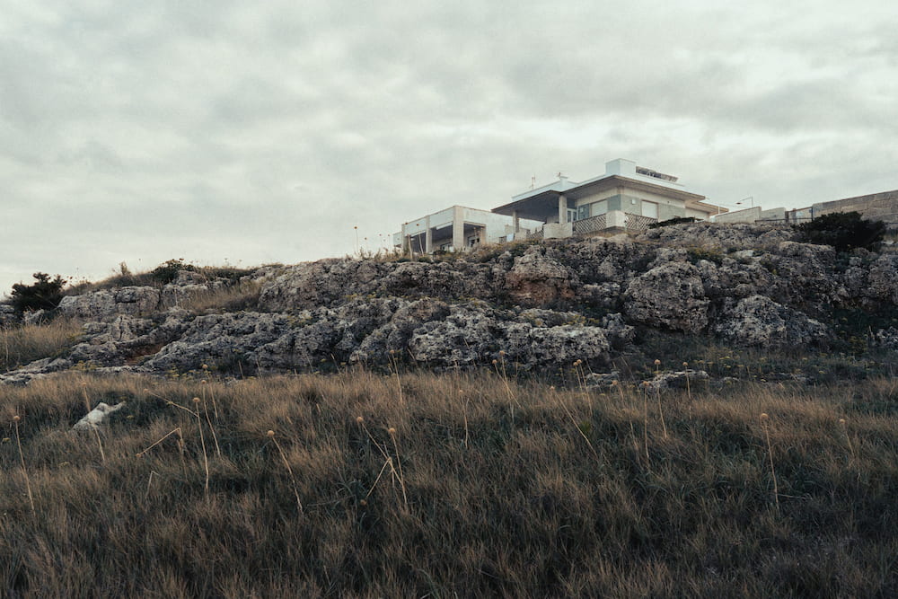 Modern white house on a ridge of grey rock above dry grass under an overcast sky