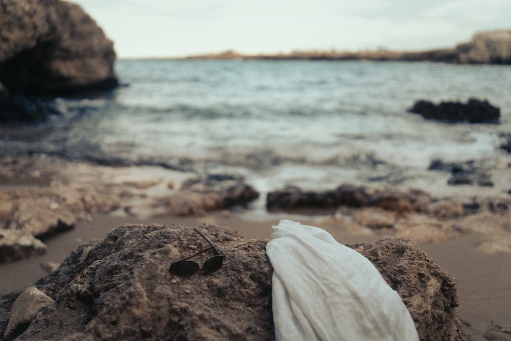 Sunglasses and a white shirt on a rock, a sandy shore and small waves blurred behind