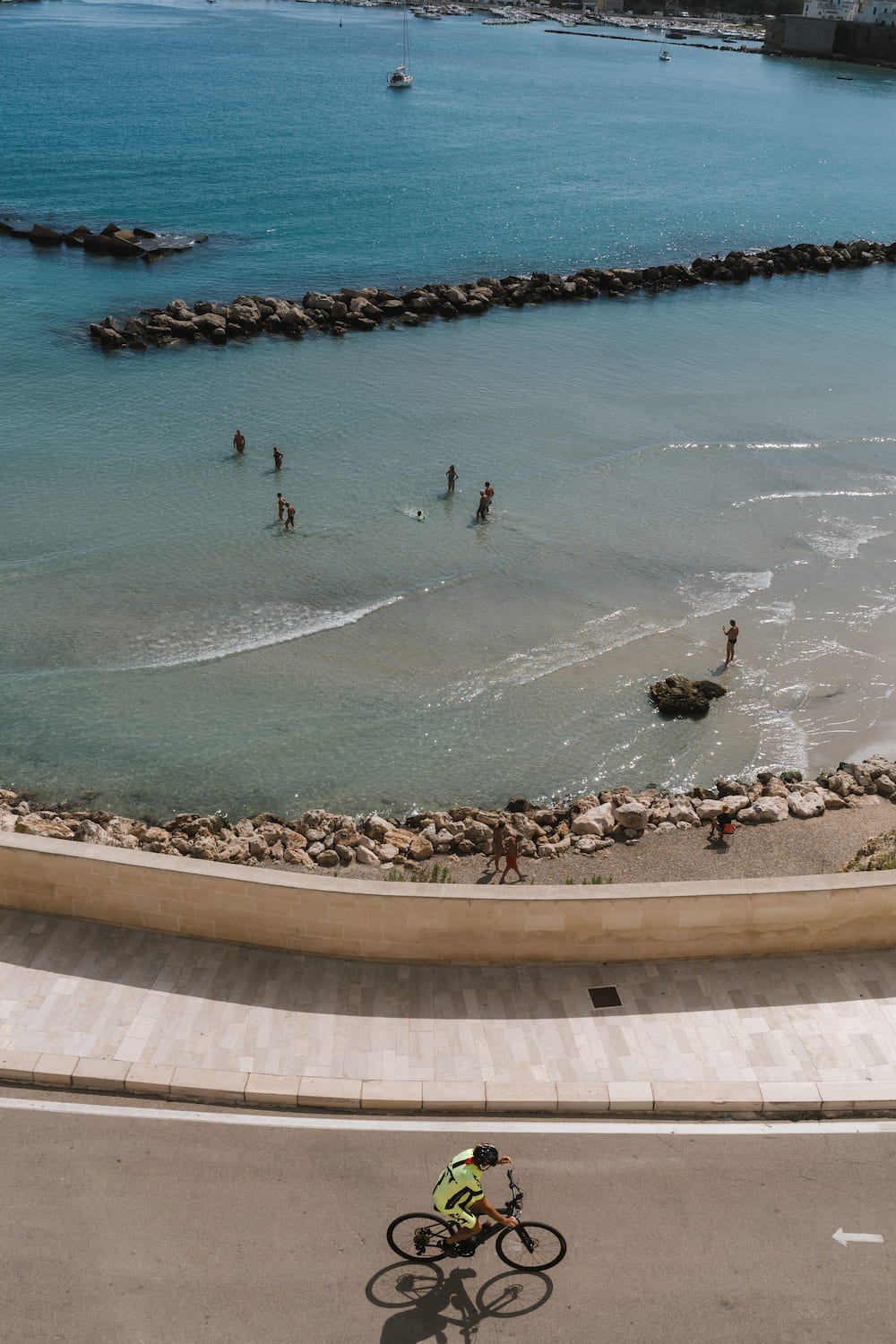 Cyclist in yellow on a seafront road above people wading in clear water behind rock breakwaters