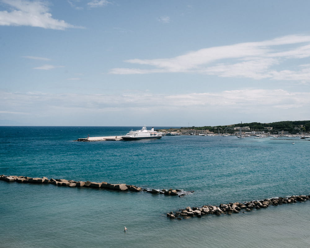 Cruise ship moored at a harbour pier beyond rock breakwaters, one swimmer in turquoise water