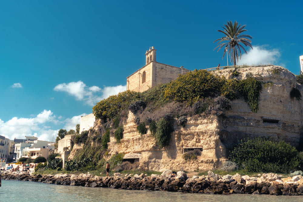 Stone church with a small bell tower and a palm on an overgrown cliff above a rocky shore