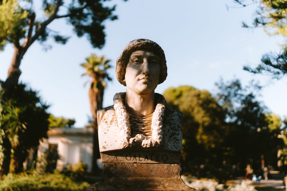 Sunlit marble bust of a man in a cap, inscribed "GALATEO", among trees and palms