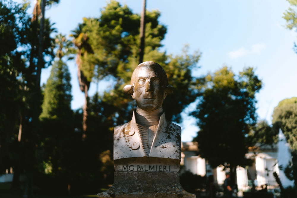 Stained marble bust of a man in a high-collared coat inscribed "MG PALMIERI" against trees and blue sky