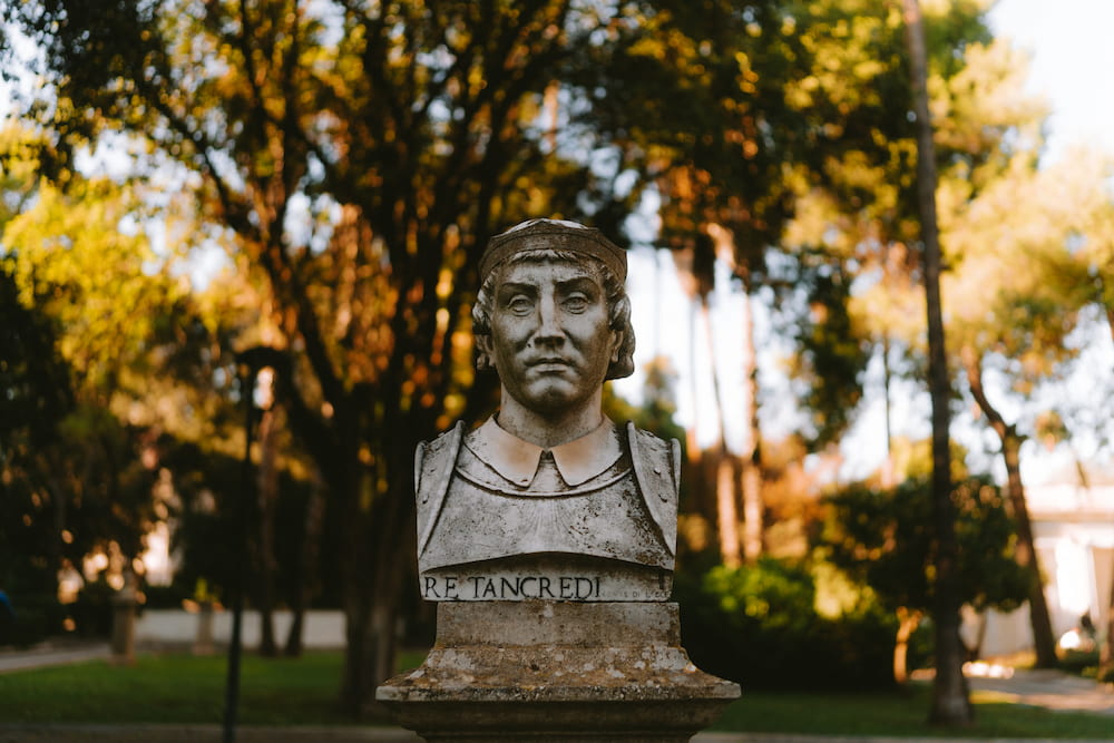 Stone bust of a man in armour inscribed "RE TANCREDI" in front of trees lit by late sun