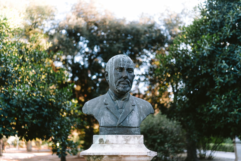 Dark bronze bust of a bearded man in a jacket on a stone plinth among green trees