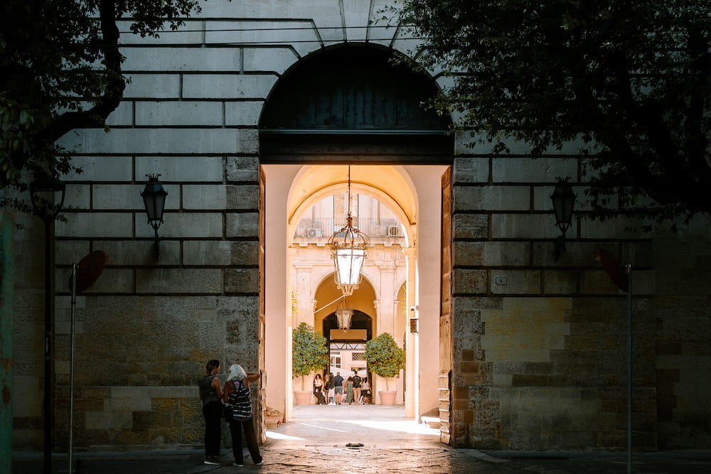 Two people at a stone gateway looking through to a sunlit arcaded courtyard with a hanging lantern