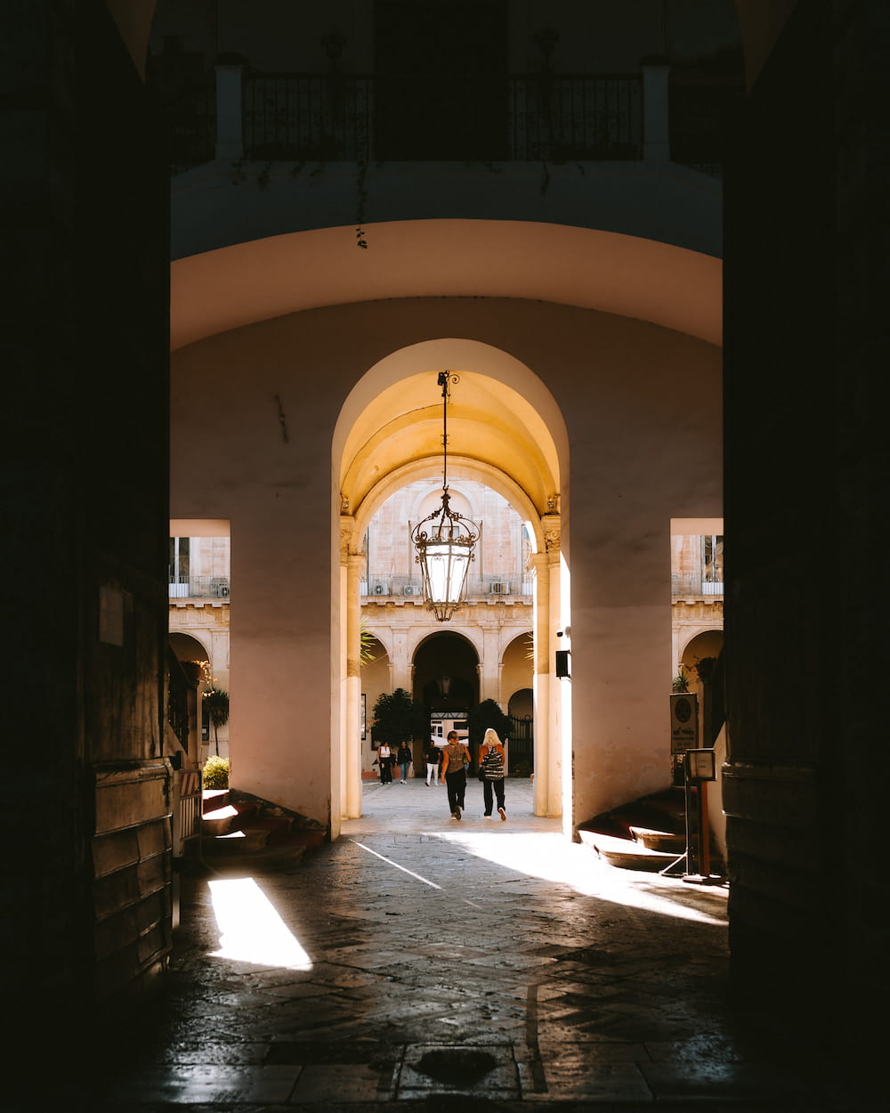 View from a dark passage through a lit arch with a hanging lantern into a busy courtyard