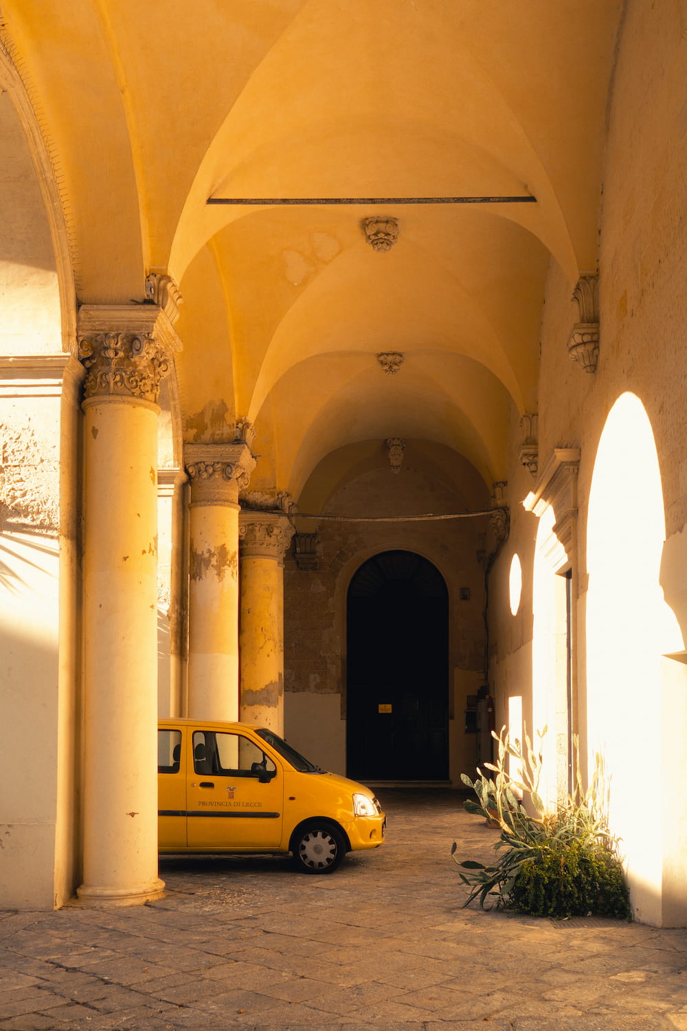Yellow car parked under a vaulted portico with stone columns in warm late sunlight