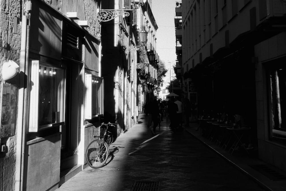 Black and white narrow street with a parked bicycle, wall lamps and people walking in shade
