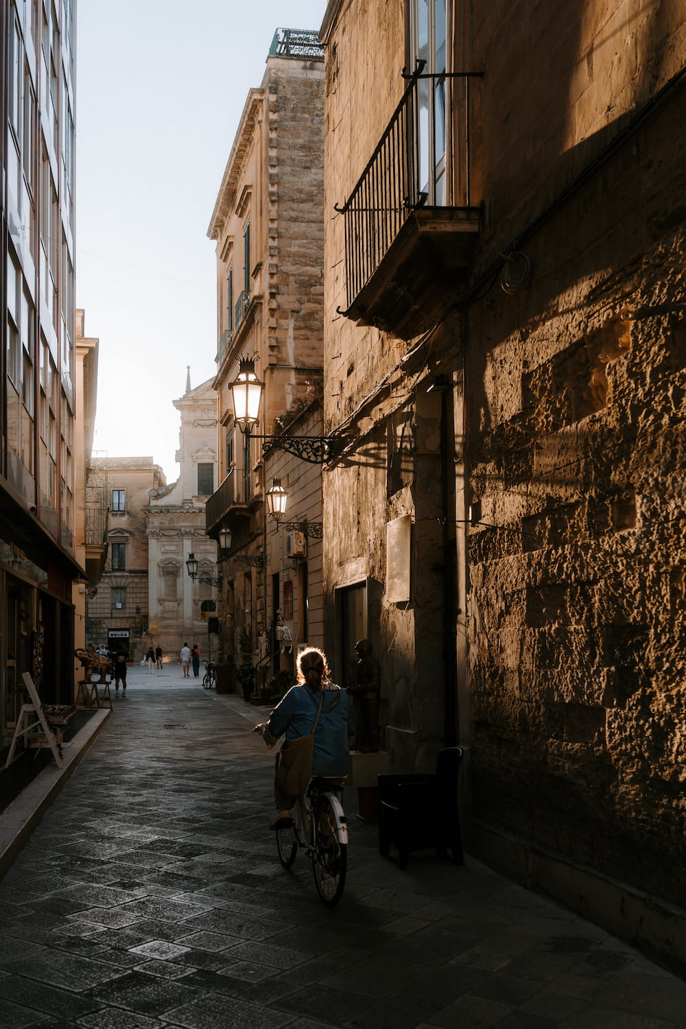 Cyclist riding down a narrow paved street between sunlit stone buildings toward a church facade