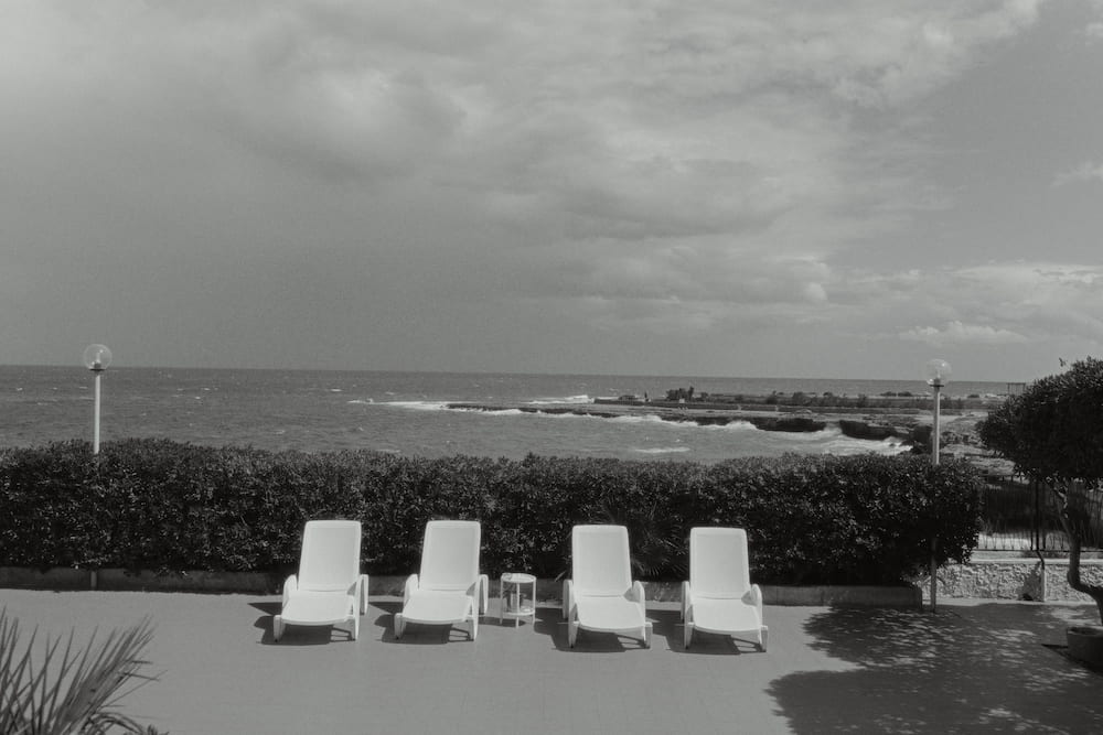 Black and white terrace with four white sun loungers before a hedge, a rough sea and heavy clouds