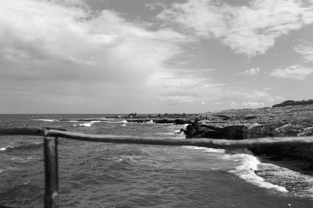 Black and white view past a wooden rail of waves breaking along a low rocky shore under clouds