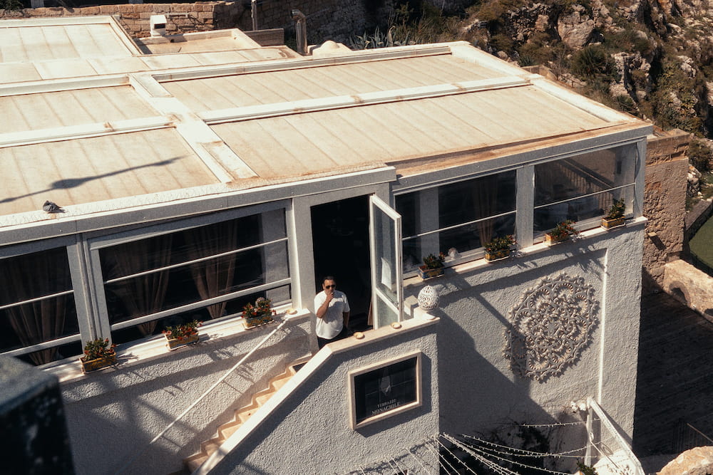 Man in sunglasses and a white T-shirt in the doorway of a glassed-in terrace, seen from above