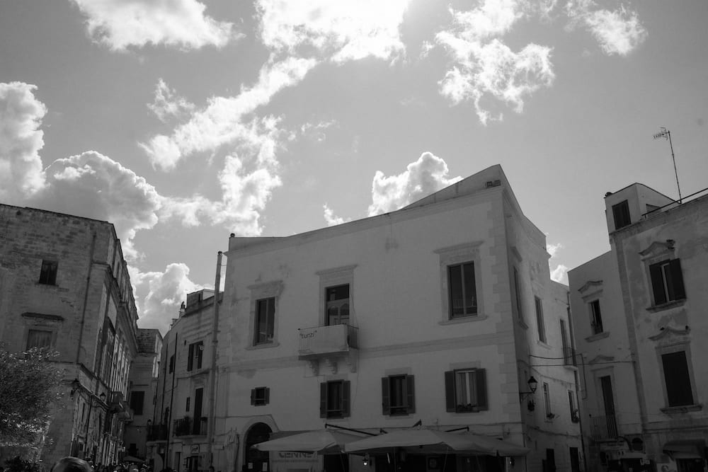 Black and white low view of pale stone buildings with shuttered windows and café umbrellas under clouds