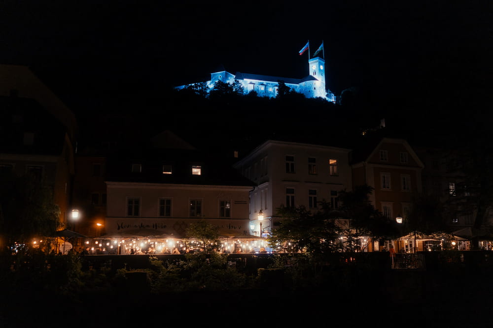 A castle lit blue on a dark hill, lit café terraces along a river below