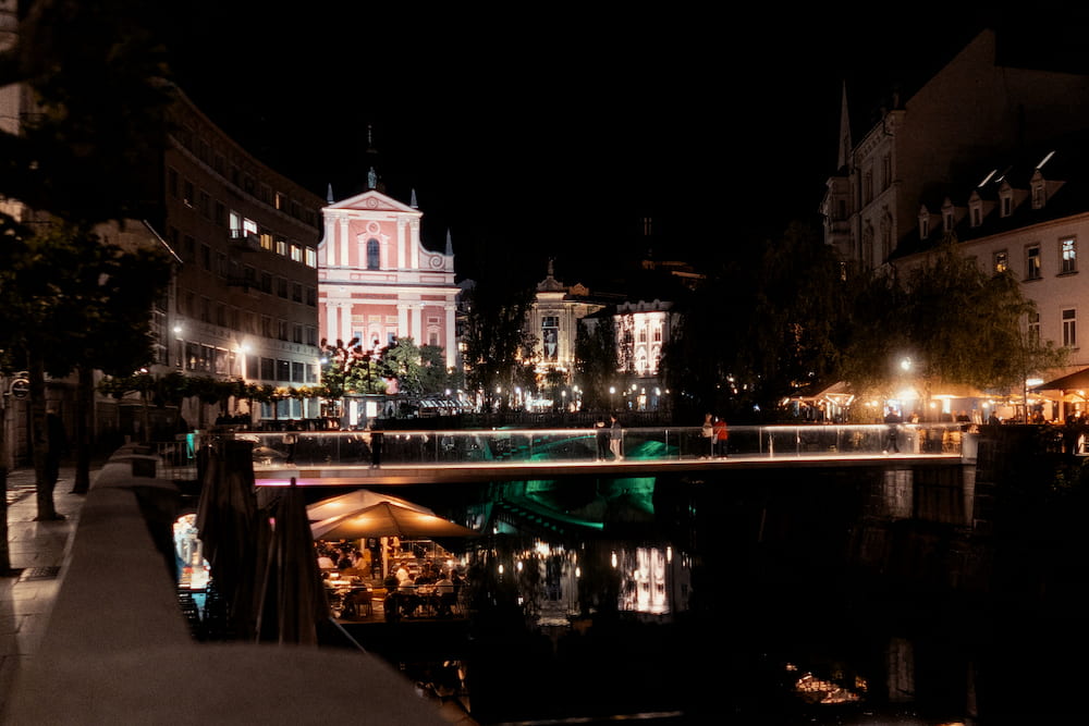 A pink church front lit at night behind a bridge, lights reflected in the river
