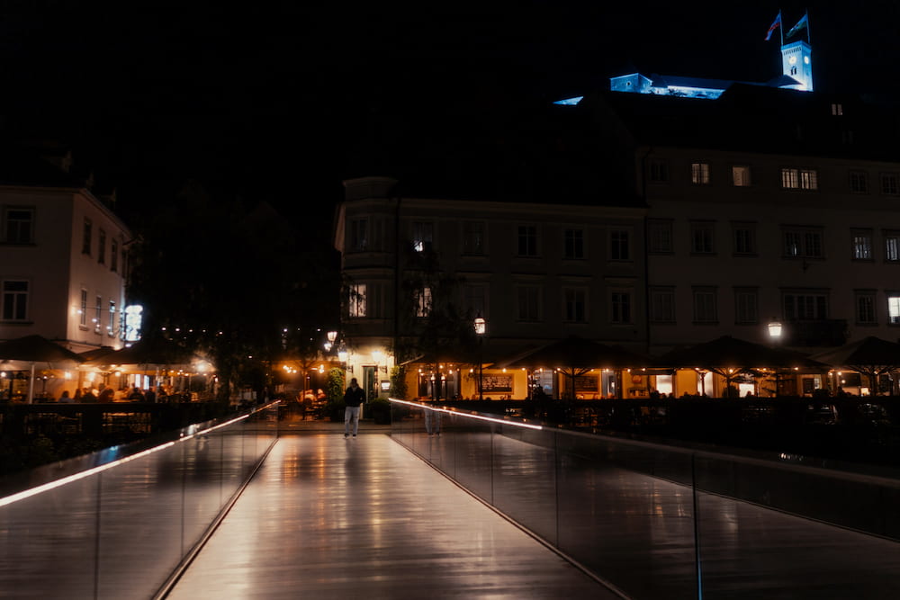 A glass-railed footbridge at night leading to lit terraces, the castle lit blue above