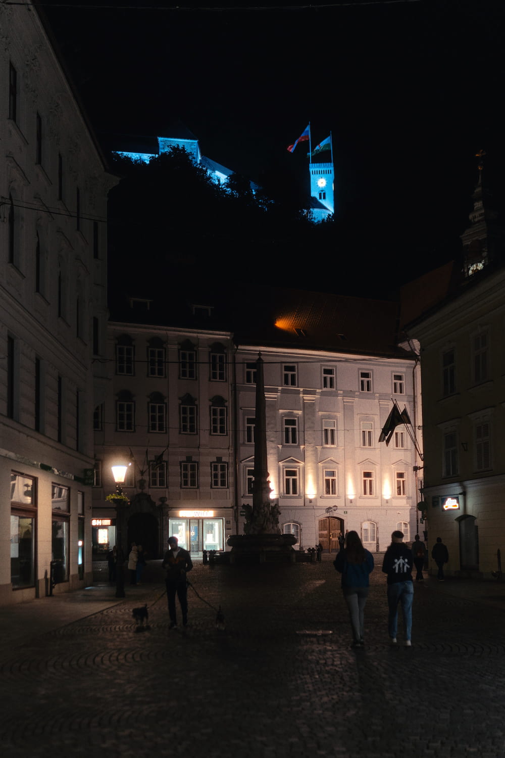 A narrow street at night, people walking, a lit blue tower at the top