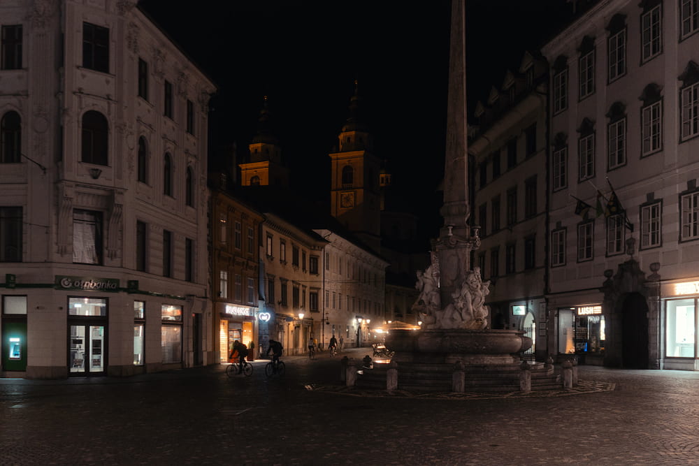 An empty square at night with a baroque fountain and an obelisk, two church towers behind