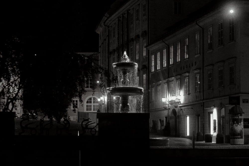 Black and white: a fountain in a square at night, lit windows behind
