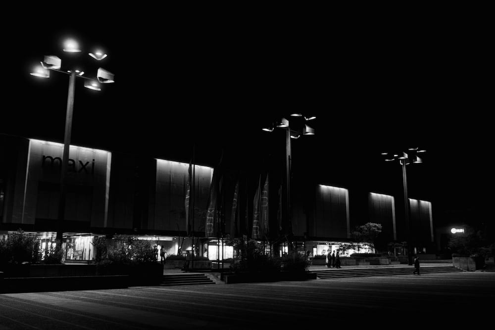 Black and white: a row of cluster street lamps along a lit building front at night