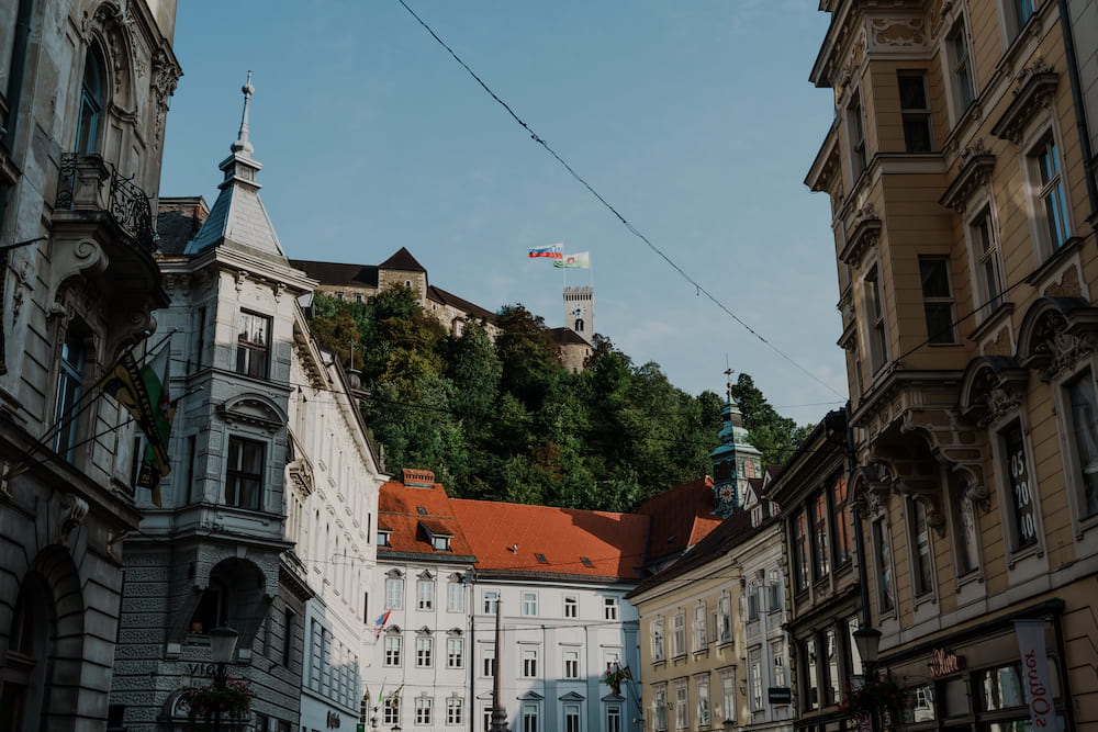 A street of old buildings with a castle on a green hill at the end, tram wires above