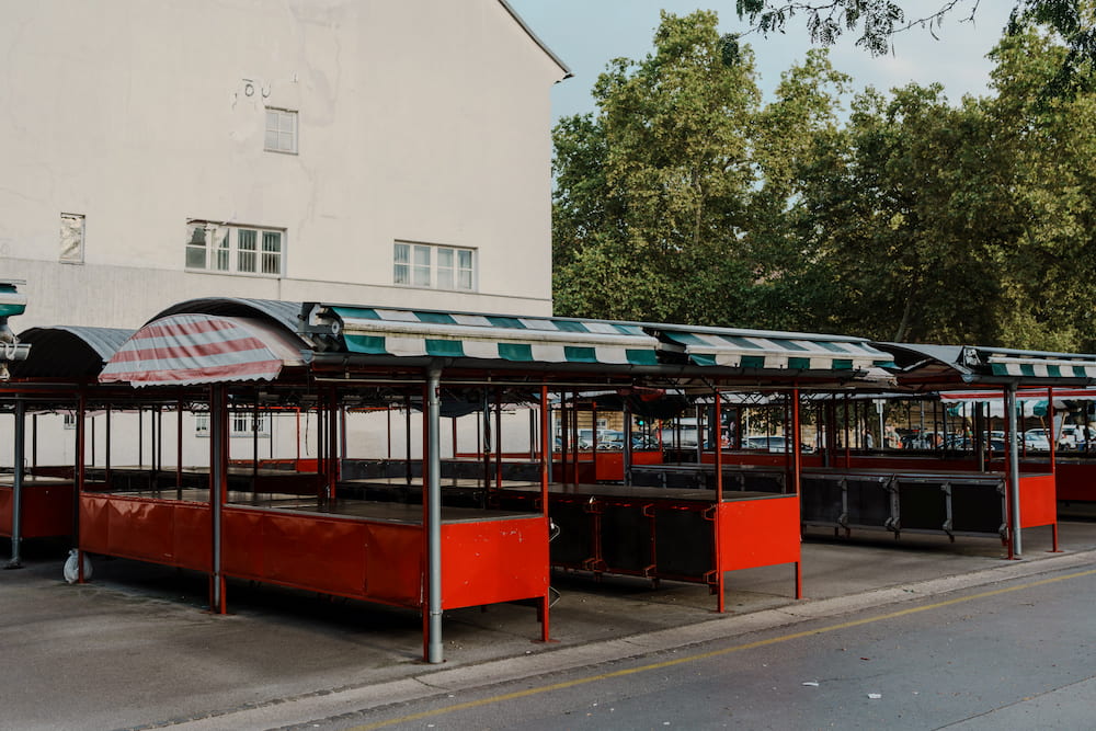 Empty red market stalls with striped awnings, trees and a white building behind