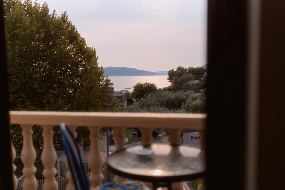 A balcony with a white balustrade and a small table, the sea and hills beyond, seen through a doorway.