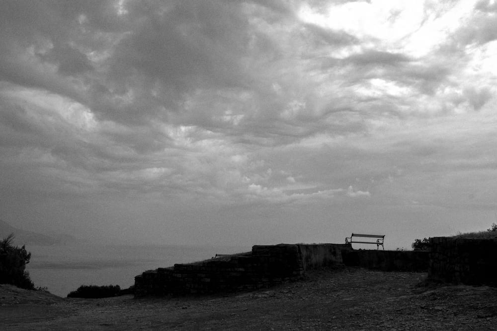 Black and white: a heavy cloudy sky over the sea, a bench on a low wall.