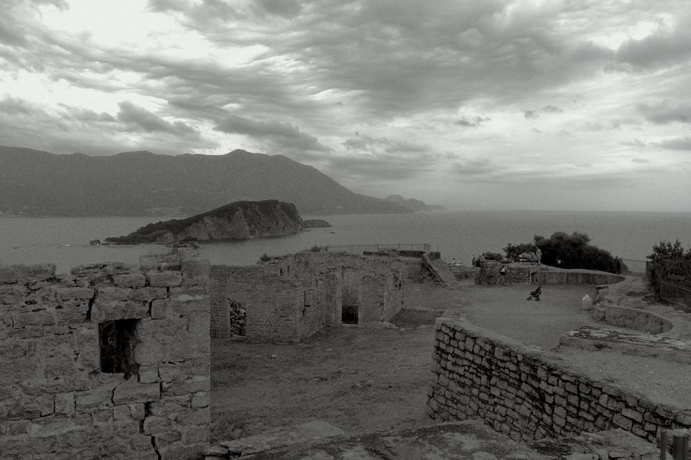 Black and white: old stone walls on a hill, an island and mountains across the water.