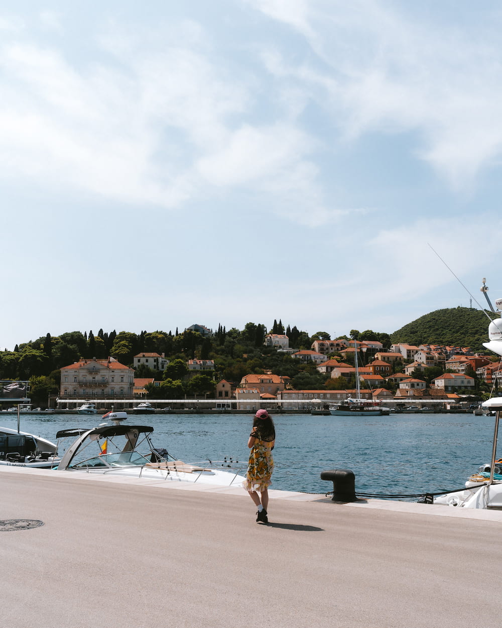 A woman walking on a harbour quay, boats and a town on the hill behind.