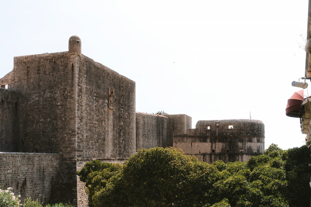 Stone city walls and a fortress above trees.
