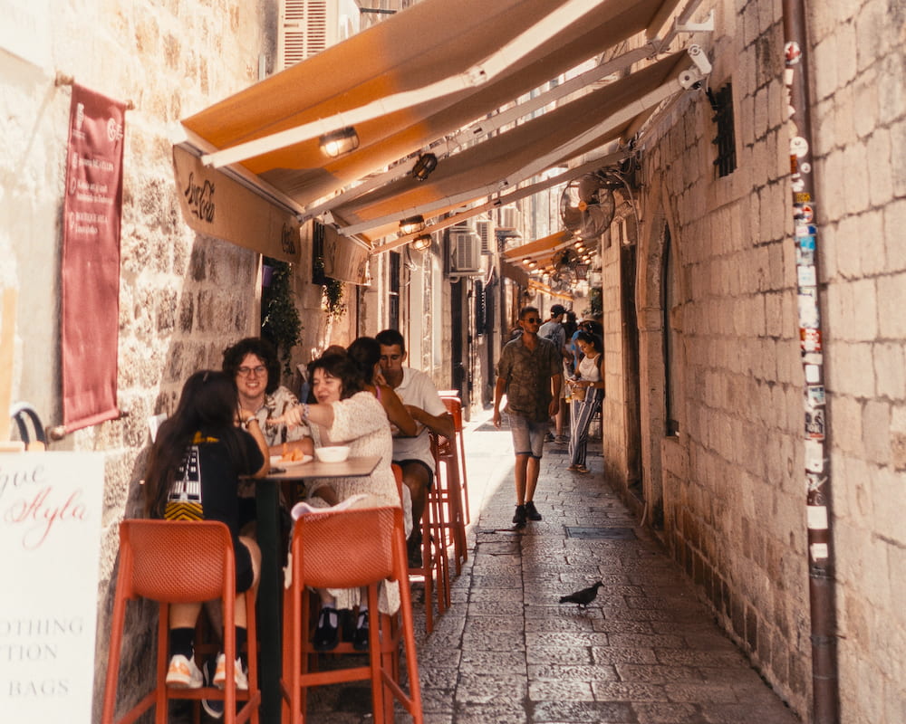 People eating at a high table under orange awnings in a narrow stone lane.